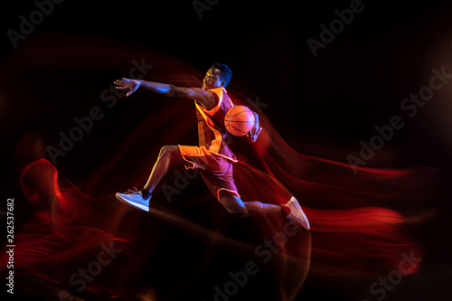 Choosing the right way. African-american young basketball player of red team in action and neon lights over dark studio background. Concept of sport, movement, energy and dynamic, healthy lifestyle.