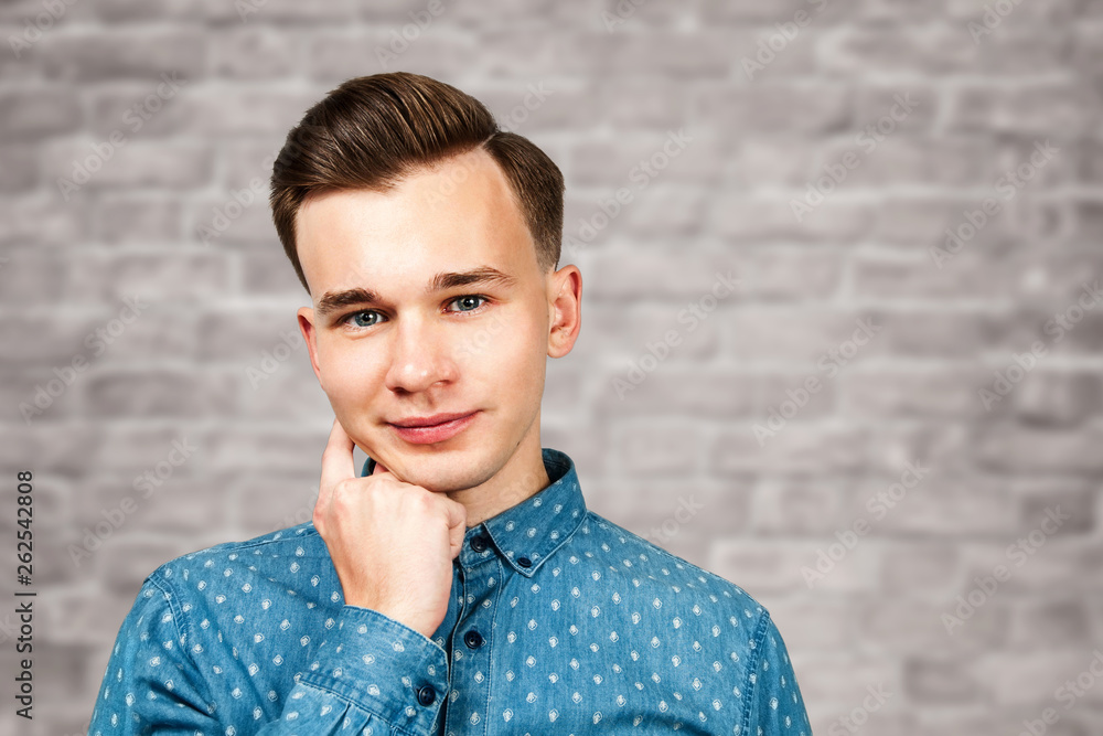 White young guy dressed in blue holds hand behind head and looking on brick wall background White young guy dressed in blue holds hand behind head and looking on brick wall background