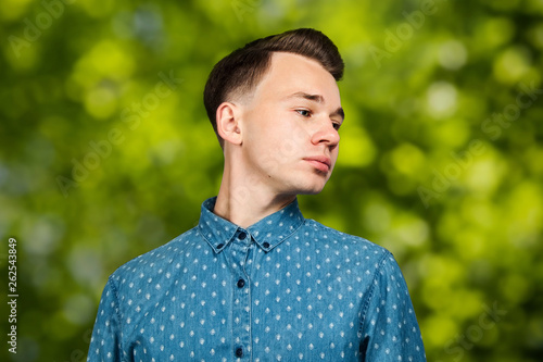 White young guy dressed in blue shirt on green bokeh background smiles and looking aside