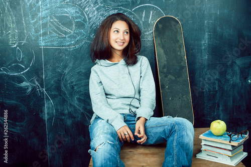 young cute teenage girl in classroom at blackboard seating on table smiling, modern hipster concept