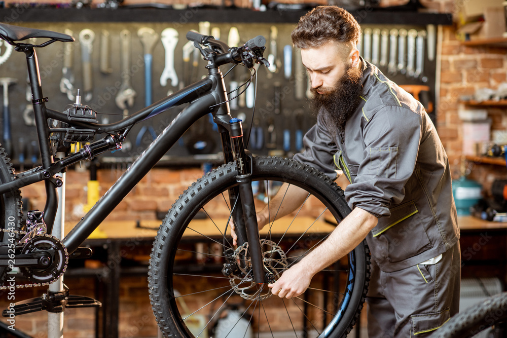 Handsome bearded repairman in workwear mounting wheel on a mountain bicycle at the workshop Handsome bearded repairman in workwear mounting wheel on a mountain bicycle at the workshop