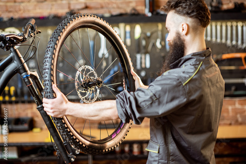Handsome repairman in workwear serving mountain bicycle  standing with front wheel at the workshop of a bicycle shop