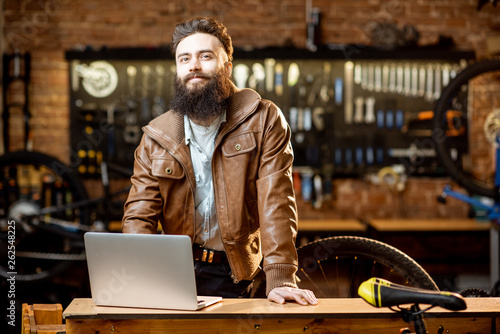 Portrait of a handsome bearded man as bicycle store owner or manager standing with laptop at the bicycle workshop