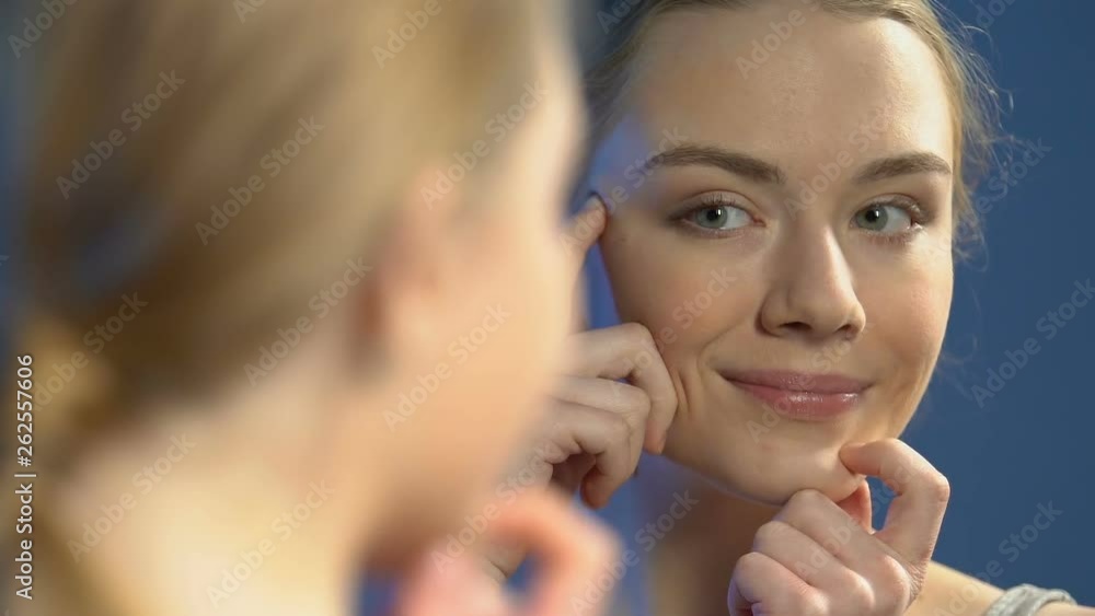 Upset female teenager looking in mirror, noticing pimples, skin imperfection Upset female teenager looking in mirror, noticing pimples, skin imperfection