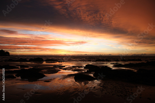 Sky with deep hanging storm clouds and wet sludge during low tide swathed in yellow and red bright light during sunset on tropical island Ko Lanta, Thailand