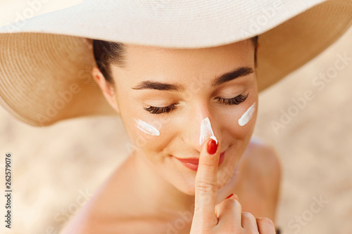 Beautiful Young woman with sun cream on face. Girl holding sunscreen bottle on the beach. Female in hat applying  moisturizing lotion on skin.Skin care. Sun protection. Suntan photo