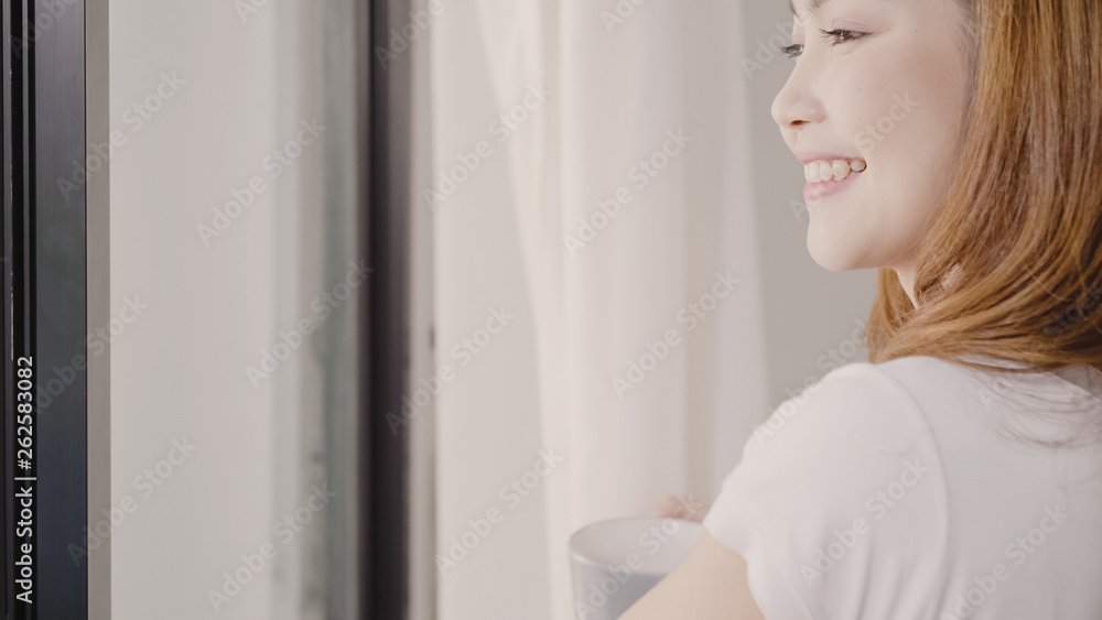 Happy beautiful Asian woman smiling and drinking a cup of coffee or tea near the window in the bedroom. Young asia female open the curtains and relax in the morning. Lifestyle woman at home concept. Happy beautiful Asian woman smiling and drinking a cup of coffee or tea near the window in the bedroom. Young asia female open the curtains and relax in the morning. Lifestyle woman at home concept.