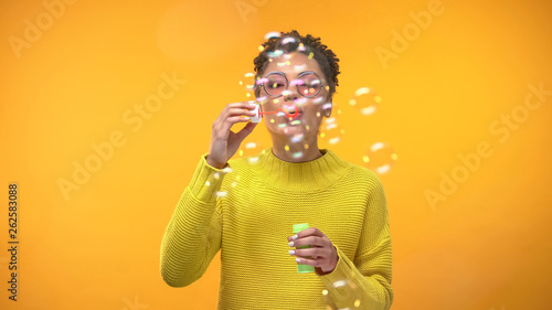 Joyful black woman blowing soap bubbles on yellow background, youth happiness