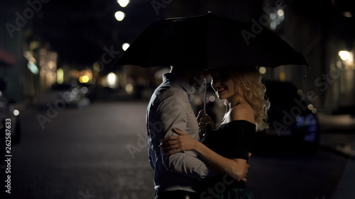 Man holding umbrella, hugging his beloved and enjoying date in the evening