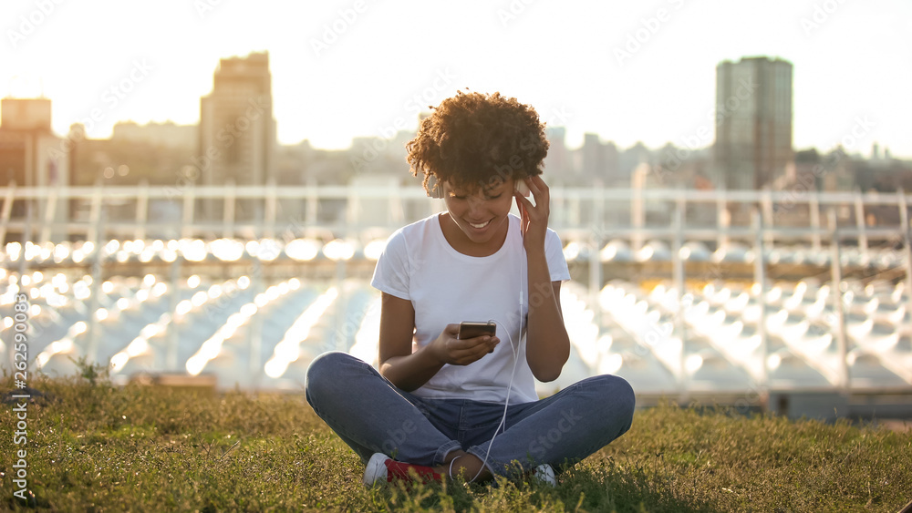 Young afro-american woman listening to music in headset, sitting on lawn, relax