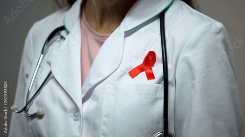 Female doctor attaching red ribbon to medical suit, AIDS awareness campaign