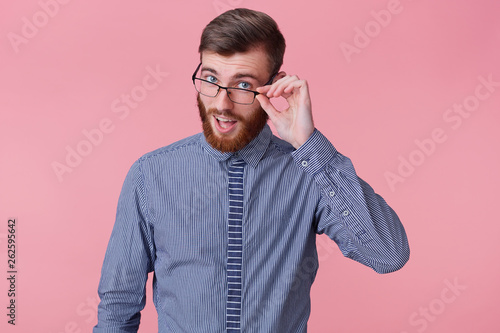 Portrait of a young handsome bearded man who tells a funny story, looks through his glasses isolated over pink background. photo