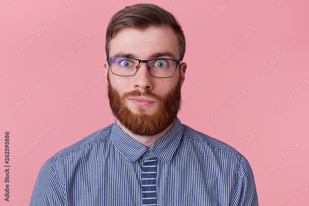 Close up portrait of funny ginger guy in glasses and striped shirt looking at camera, isolated on pink background Close up portrait of funny ginger guy in glasses and striped shirt looking at camera, isolated on pink background