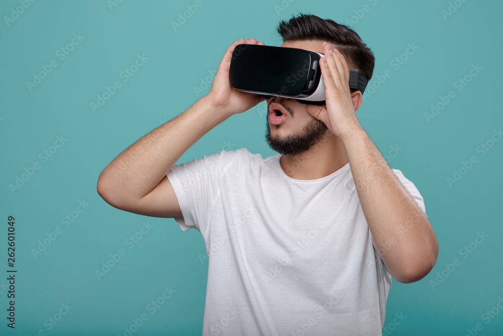 Shocked handsome bearded man wearing virtual reality goggles in studio over cyan background. Smartphone using with VR headset. Shocked handsome bearded man wearing virtual reality goggles in studio over cyan background. Smartphone using with VR headset.