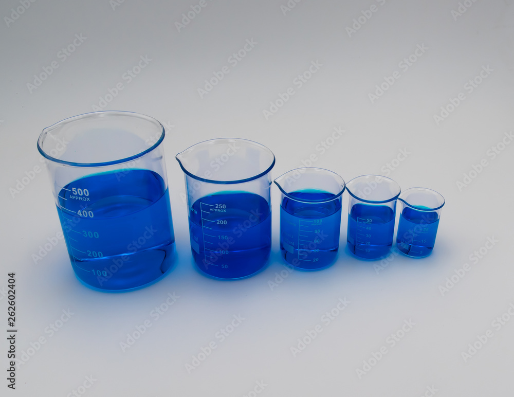 Five round chemistry beakers filled with blue liquid in a line across a white background with natural shadows Five round chemistry beakers filled with blue liquid in a line across a white background with natural shadows