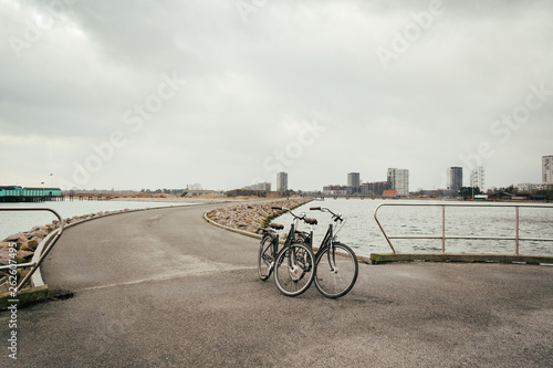 Two identical retro black city bicycles parked on embankment, road along Baltic Sea, embankment with asphalt path background of sdan and city Copenhagen Denmark in winter cloudy weather