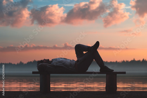 A man is lying on a wooden bench in a picturesque place. Lake and forest in the background.
