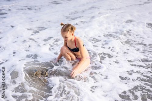 Having fun and joy concept. Blonde lovely girl with two braids playing with sea sand. Playful active kid on beach with waves in summer vacation