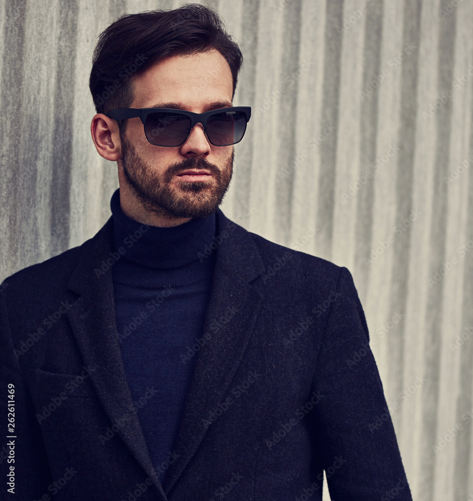 Fototapeta premium Fashion beard style business handsome male model in fashion sunglasses and blue coat with serious concentrated look posing in style clothing on wall outdoors background. Closeup