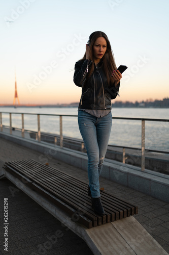 Fototapeta Naklejka Na Ścianę i Meble -  Young woman listens to music in closed headphones through her phone wearing a leather jacket and jeans at a sunset near river Daugava