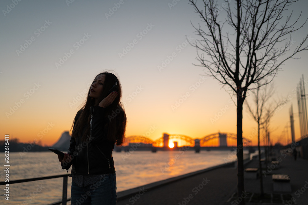 Young woman listens to music in closed headphones through her phone wearing a leather jacket and jeans at a sunset near river Daugava