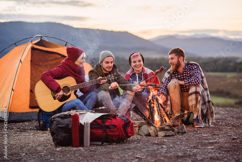 Happy friends having a picnic at the mountains, they are chatting, drinking energy drinks, playing on guitar and bake sausages on campfire. Concept of happiness, youth and enjoyment of life.