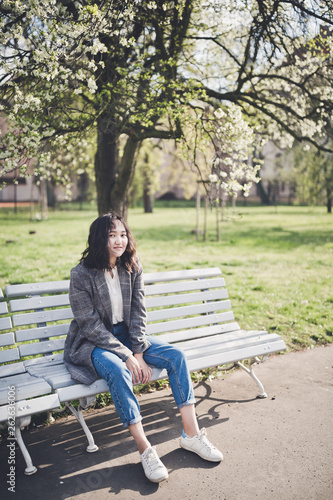 Cheerful asian young woman sitting on a bench under the blooming trees in a park © lermont51