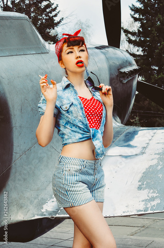 Pin up beautiful girl with red hair stands against the background of an old plane