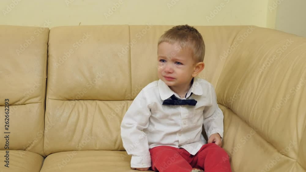 Elegant baby child sitting on sofa and crying, lonely kid with bow Elegant baby child sitting on sofa and crying, lonely kid with bow