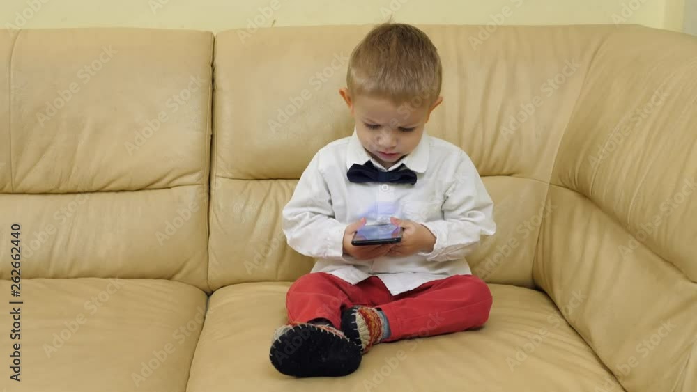 Portrait of fashionable child with boy, looking at mobile phone, little businessman on sofa, modern enterteinment Portrait of fashionable child with boy, looking at mobile phone, little businessman on sofa, modern enterteinment