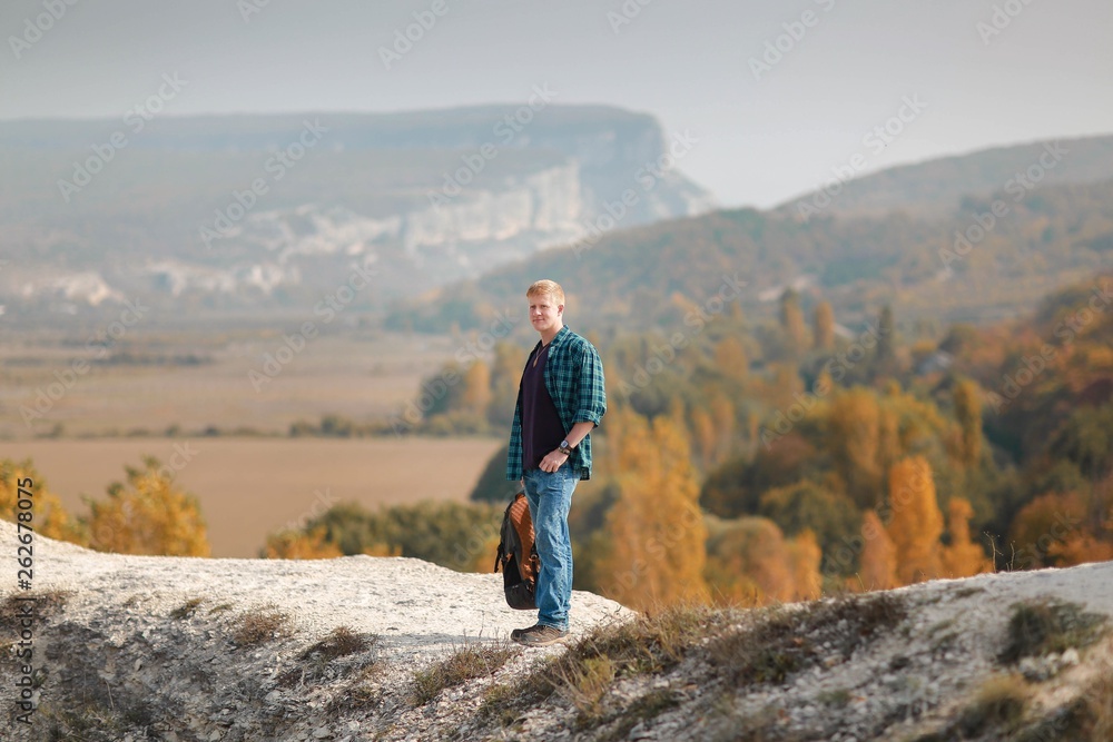 Redhead man in a plaid shirt and purple t-shirt with a backpack in his hand stands on a mountain trail in the Crimean mountains in autumn. Hiking and adventure concept.