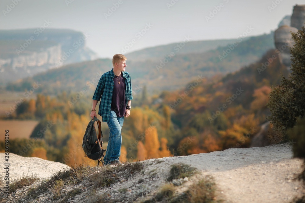 Redhead man in a plaid shirt and purple t-shirt with a backpack in his hand goes along a mountain trail in the Crimean mountains in autumn. Hiking and adventure concept.