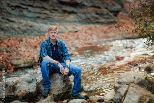 Redhead man in a plaid shirt and purple t-shirt sits on a stone in the autumn forest near a mountain stream in the Grand Canyon of Crimea. Autumn, travel and hiking concept.
