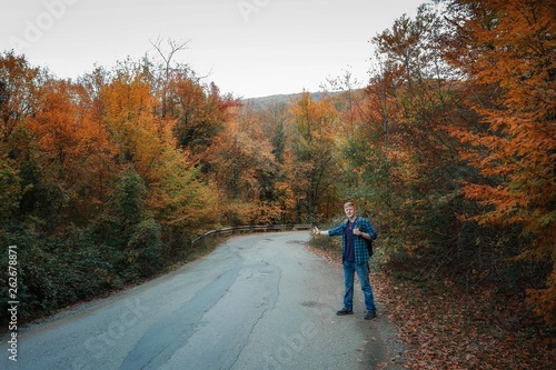 Redhead man in a plaid shirt and purple t-shirt with backpack hitchhikes on the road in the middle of the autumn forest. Autumn, travel and tourism concept.