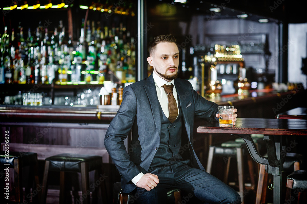 serious man in a stylish suit drinking whiskey in a pub. Stylish handsome male in an elegant suit holds a glass of whiskey at bar counter background.