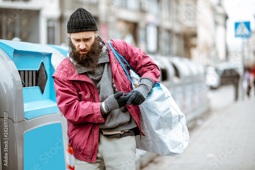 Portrait of depressed homless beggar standing with bag near the trash containers in the city photo