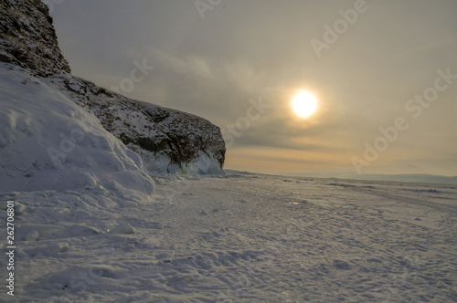 Rock on the shore of a snow-covered lake in the rays of sunset © Евгений Донец
