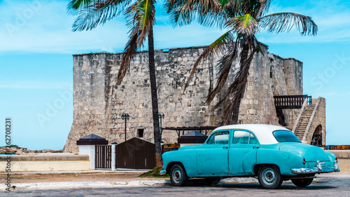 Fototapeta Naklejka Na Ścianę i Meble -  Havana, Cuba. Under the palm tree an old American car parked at it`s shade, besides of an old fort.
