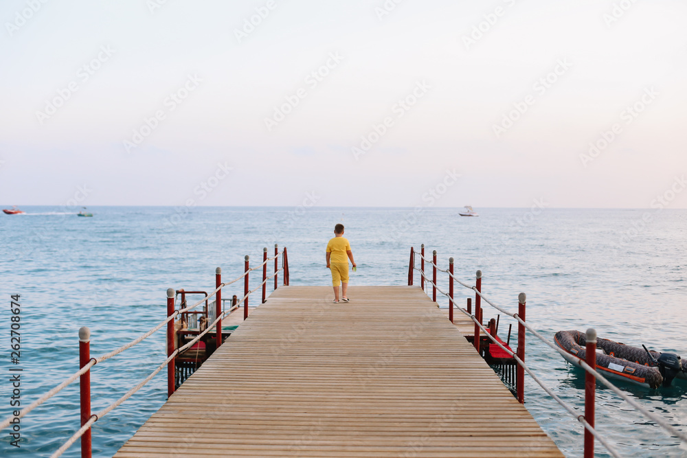Naklejka premium Boy walking on the pier and looking at the sea horizon and blue sky. Vacation at sea. Summer time sea vacation background 