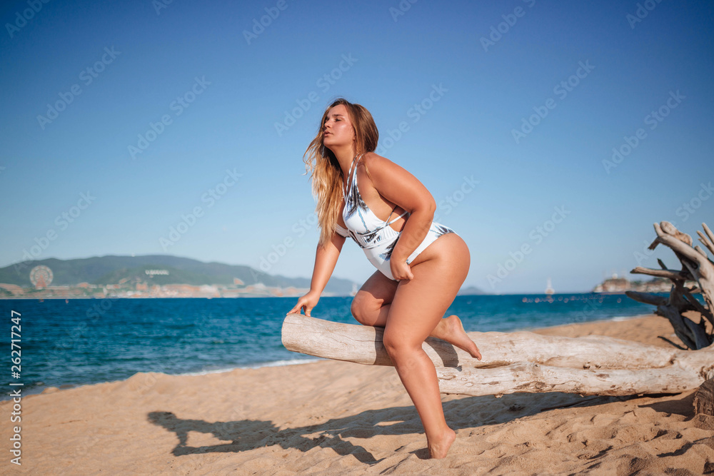 Overweight girl, beautiful young girl in a white one-piece swimsuit on the beach at sea on white sand with a big log posing. Horizontal photo close-up portrait