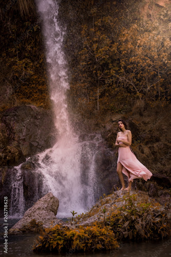 Obraz premium Attractive girl posing on the rock among in fairy fast forest in autumn and beside beautiful waterfall with blue water. Fairy tale.