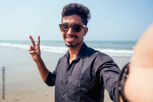 young hindu stylish man taking a picture self portrait on the smartphone front camera with sunglasses active beach vacation on semmertime happy Goa India beach. sunscreen spf protection concept.