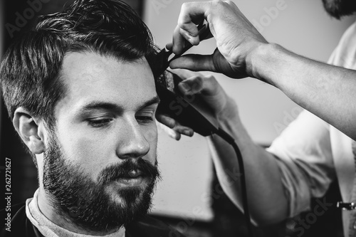 Young handsome barber making haircut for attractive bearded man at barbershop. Black and white or colorless photo. Hairstyle, salon, hairdresser, barber shop, lifestyle concept. Caucasian male models.