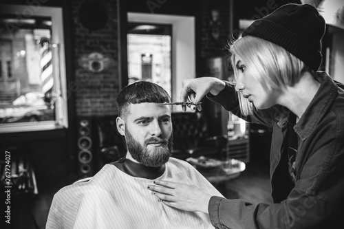 Young woman making haircut for bearded man at barbershop. Female barber at salon. Gender equality. Woman in the male profession. Black and white or colorless photo. Hairstyle, salon, hairdresser