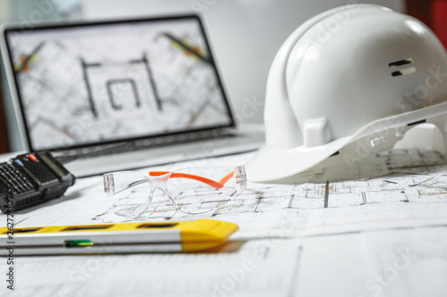 White hard hat, laptop with drawings, glasses and walkie talkie with and blueprints on a table