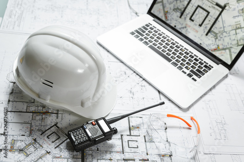 White hard hat, laptop with drawings, glasses and walkie talkie with and blueprints on a table