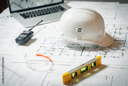 White hard hat, laptop with drawings, glasses and walkie talkie with and blueprints on a table