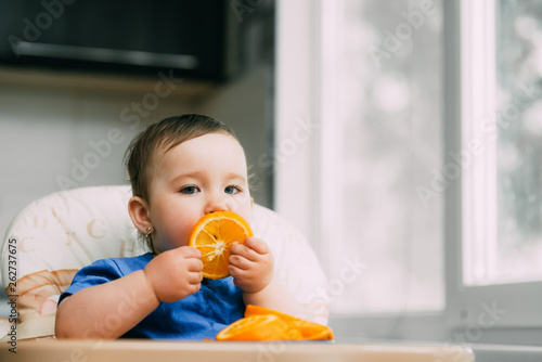 a little girl in a blue t-shirt and a blue plate sitting in a child's chair eating an orange