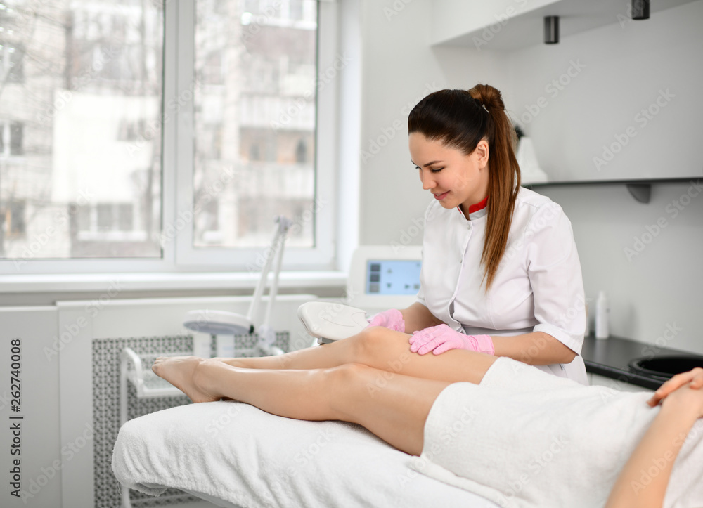 Doctor-cosmetician brunette performs a cosmetic procedure on the patient's legs with electronic medical device in a bright beauty shop Doctor-cosmetician brunette performs a cosmetic procedure on the patient's legs with electronic medical device in a bright beauty shop
