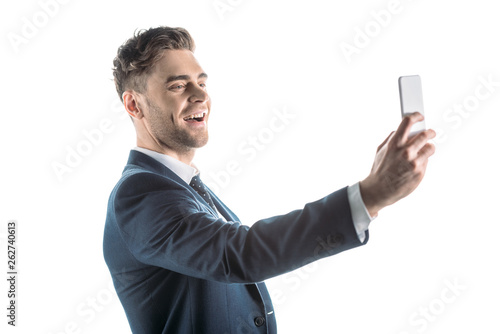 handsome, cheerful businessman taking selfie with smartphone isolated on white